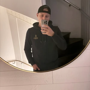 Svart hoodie från Morris - Svart hoodie från Morris med guldfärgad brodyr på bröstet och snörning i huvan. Klassisk passform med långa ärmar och ribbade muddar. Perfekt för dig som gillar stilrena och bekväma plagg med snygga detaljer.