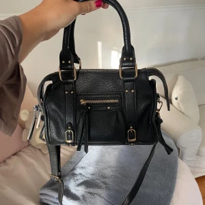 Svart handväska med gulddetaljer - Snygg svart handväska (fakeskinn) köpt i Spanien så oklart vilket märke det är. Som ny skick 