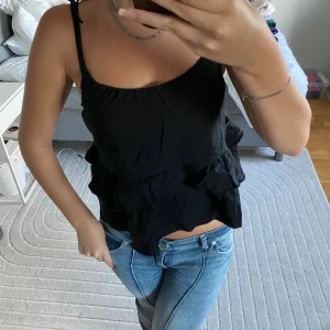 Svart volangtop med knytband - Supersöt svart linnetopp med volanger och tunna axelband som knyts upptill. Toppen har en luftig och flowy passform, perfekt för varma dagar. Materialet känns lätt och toppen har en feminin vibe med sina dubbla volanglager.