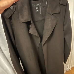 Svart trenchcoat från New Look - Stilren svart trenchcoat i mocka tyg från New Look med klassisk krage och bälte i midjan. Jackan har långa ärmar och en rak, loose passform som ger en cool och avslappnad vibe. Perfekt för höst och vår när du vill ha en snygg och enkel look.