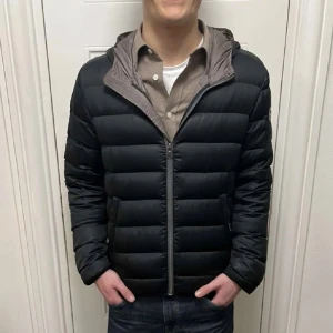 Svart dunjacka från Colmar med huva - Svart dunjacka från Colmar med huva och dragkedja framtill. Jackan har quiltad design, två fickor med dragkedja och en diskret logotyp på ärmen. Perfekt för kyliga dagar och har en stilren, sportig look. Ena framfickan går inte att stänga.