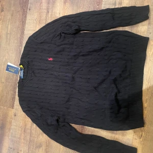 Svart stickad tröja från Polo Ralph Lauren - Kabelstickad svart tröja från Polo Ralph Lauren med klassisk rund halsringning och röd broderad logga på bröstet. Tröjan har långa ärmar och ribbade muddar vid ärmslut och nederkant. Perfekt för lager-på-lager under kyliga dagar.