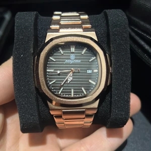 Roséguld klocka  - Stilren klocka från Pagomu i roséguld med länkarmband i metall. Urtavlan är svart med horisontella ränder och har datumvisning. Boetten är åttakantig och detaljerna på urtavlan är i matchande roséguld. Perfekt accessoar för dig som gillar snygga detaljer.