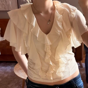 Volangblus i ljusbeige från Kappahl - Supersöt ljusbeige blus från Kappahl med v-ringning och stora volanger framtill och på ärmarna. Blusen är tunn och luftig med korta, vida ärmar och figurnära passform. Perfekt för vår och sommar när du vill ha något extra fint.