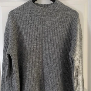 Grå ribbstickad tröja från H&M - Mysig grå ribbstickad tröja från H&M med hög hals och långa ärmar. Tröjan har en rak passform och slits i sidorna för en snygg detalj. Perfekt för lager-på-lager under kyliga dagar.