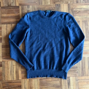 Mörkblå merinoullströja J.Lindeberg - Tröjan har krympt från L till XS. Snygg mörkblå stickad tröja från J.Lindeberg i 100% merinoull. Klassisk rund halsringning och ribbade muddar vid ärmslut och nederkant. Perfekt för dig som gillar stilrena och bekväma plagg med premiumkänsla.