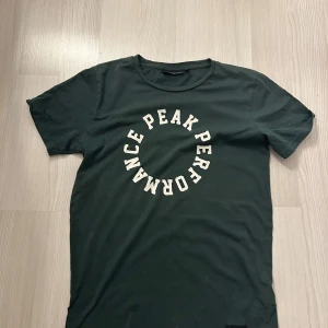 Mörkgrön Peak Performance t-shirt - Snygg mörkgrön t-shirt från Peak Performance med vit tryckt logga i cirkelform på bröstet. Klassisk rund hals och korta ärmar. Tillverkad i mjuk bomull som är skön att bära hela dagen.