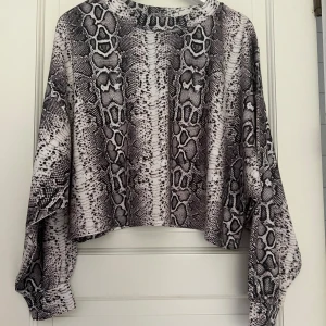 Croppad tröja med ormmönster - Säljer en croppad långärmad tröja med ormmönster. Tröjan har vida ärmar med resår vid ärmsluten och en avslappnad passform.  Helt ny utan prislapp.
