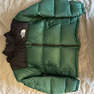 Grön The North Face pufferjacka S - Säljer en grön och svart pufferjacka från The North Face i storlek S. Jackan har en klassisk boxig passform, hög krage och dragkedja framtill. Fylld med dun och har två sidofickor samt logga på bröstet och axeln. Perfekt för kalla dagar.