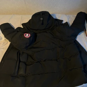 Svart dunjacka från Canada Goose - Säljer en svart dunjacka från Canada Goose med klassisk patch på ärmen. Jackan har hög krage, dragkedja och medföljande pälskrage. Perfekt för kalla vinterdagar och riktigt snygg streetstil.