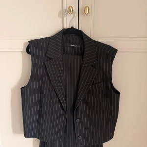 Randig svart väst från Gina Tricot - Snygg svart väst med vita tunna ränder från Gina Tricot. Klassisk kavajkrage, knäppning med två knappar framtill och markerade axlar. Perfekt för en trendig och stilren look. Västen har en rak passform och är tillverkad i ett mjukt polyestertyg. Kommer med likadana byxor, byxorna i 36 och västan i 34.