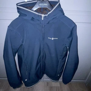 Marinblå vindjacka från Sail Racing 160 - Snygg marinblå vindjacka från Sail Racing med huva och dragkedja framtill. Jackan är i väldigt bra skick. Logga på bröstet och reflekterande detalj på huvan. Insidan är fodrad med mjuk fleece för extra komfort.