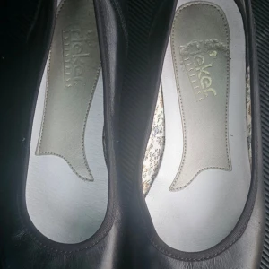 Svarta ballerinaskor med klack från Rieker - Snygga svarta ballerinaskor från Rieker med låg klack och rund tå. Utsidan är i mjukt skinn och insidan har ett blommigt mönster som ger extra stilpoäng. Sulan är beige och har bra grepp. Perfekt för dig som gillar klassisk look med en twist.