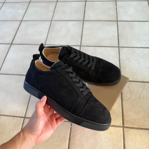 Svarta sneakers från Christian Louboutin - Snygga svarta sneakers i mocka från Christian Louboutin med klassisk röd sula och diskret logga undertill. Skorna har rund tå, svarta snören och en stilren design som passar till det mesta. Insidan är ljusbeige och sulan är platt för en clean look.