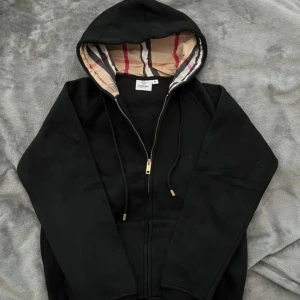 Svart Burberry hoodie med dragkedja - Svart hoodie från Burberry med klassiskt rutigt foder i huvan. Tröjan har dragkedja framtill, snörning vid huvan och känguruficka. Den är i storleken M men den är liten i storleken så passar bättre på en S. Den är aldrig använd utan endast testad!😊