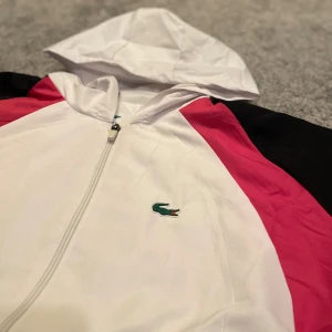 Lacoste vit och rosa vindjacka - Snygg vindjacka från Lacoste med huva, dragkedja och stora loggan på ryggen. Jackan är vit med rosa paneler och svarta ärmar, tillverkad i lätt polyester. Perfekt för dig som gillar sportig och clean stil. Klassisk krokodillogo på bröstet.