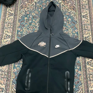 Nike svart och grå vindjacka med huva - Nike vindjacka i svart och mörkgrått med hel dragkedja och huva. Jackan har två fickor med dragkedja och vita loggor på bröstet. Snygg kontrast mellan färgerna och coolt snitt över axlarna. Perfekt för dig som gillar sportig streetstyle.