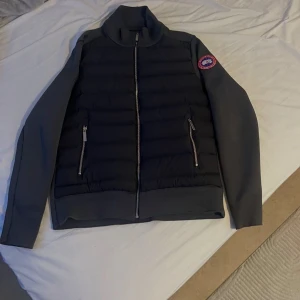 Grå cardigan från Canada Goose - Säljer en grå cardigan  från Canada Goose med quiltad framsida och två dragkedjefickor. Jackan har hög krage, långärmade släta ärmar och klassisk logga på axeln. Perfekt för kyliga dagar och riktigt snygg till streetwear.