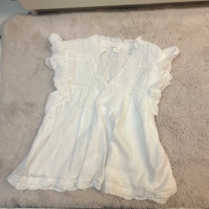 Vit blus med volang från Gina Tricot - Supersöt vit blus från Gina Tricot med volangdetaljer längs ärmarna och spetsinslag. Blusen har v-ringning och är ärmlös med romantisk känsla. Perfekt för varma dagar och enkel att matcha med jeans eller kjol.