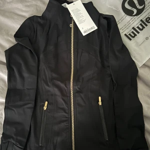 Svart Define Jacket från Lululemon - Säljer en svart Define Jacket från Lululemon med snygga guldiga dragkedjor och figurnära passform. Jackan har två fickor med dragkedja och hög krage. Perfekt för träning eller som en sportig vardagsjacka. Materialet är mjukt och stretchigt för maximal komfort.