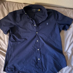 Blå kortärmad skjorta från Ralph Lauren - Säljer en stilren blå skjorta från Ralph Lauren i modellen Custom Fit. Skjortan har korta ärmar, klassisk krage och vita knappar framtill. Materialet är mjuk bomull och den ikoniska loggan är broderad på bröstet. Perfekt för dig som gillar klassisk och clean stil. Ganska tajt för att vara XL sitter mer som en L.