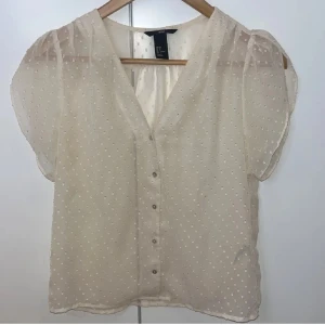 Prickig transparent blus från H&M - Säljer en söt och luftig blus från H&M i storlek 36. Blusen är transparent, beige med små vita prickar, har korta puffärmar och v-ringning med knappar framtill. Perfekt för dig som gillar romantisk och feminin stil. 😊💕 Passar till alla tillfällen.