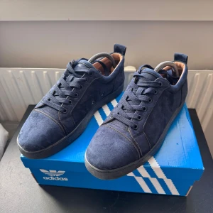 Mörkblå mockasneakers från Louboutin - Säljer ett par mörkblå sneakers från Christian Louboutin i mjuk mocka med klassisk röd sula. Skorna har rund tå, snörning och beige skinnfoder på insidan. Perfekta för dig som vill ha en stilren och lyxig sneaker med ikoniska detaljer. Priset går att diskutera 