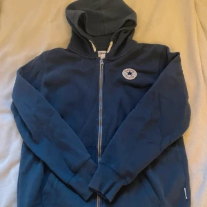 Blå hoodie från Converse med dragkedja - Mörkblå hoodie från Converse med huva och dragkedja framtill. Tröjan har ett klassiskt Converse-märke på bröstet, ribbade muddar och fickor på sidorna. Perfekt för chill dagar och enkel att matcha med jeans eller joggers. För ungdomar men passar mig bra som är S/M 