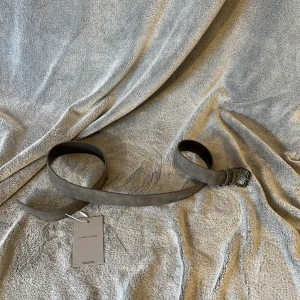 Grått mockabälte - Stilrent mörkgrått bälte i tunt lager mocka med klassiskt svart metallspänne. Bältet har en enkel och clean design utan mönster, perfekt för att lyfta vilken outfit som helst. Bältet är rakt och har flera hål för justerbar passform. Längd ~110 cm 