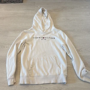 Vit hoodie från Tommy Hilfiger - Vit hoodie från Tommy Hilfiger med broderad logga och text 'EST. 1985' på bröstet. Klassisk känguruficka framtill, ribbade muddar och huva. Mjuk insida i bomullsmix, perfekt för chill dagar. Diskret Hilfiger-flagga på ärmslutet för extra stil.