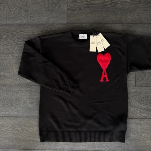 Svart Ami Paris tröja  - Svart stickad tröja från Ami Alexandre Mattiussi med ett stort rött hjärta och bokstaven A på bröstet. Rund halsringning och ribbade muddar vid ärmslut och nederkant. Perfekt för dig som gillar stilrena detaljer och vill sticka ut med en ikonisk design.