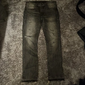 Undercover jeans - Säljer ett par grå  jeans från Undercoverism med slitna detaljer, trasiga partier och råa benslut. Jeansen har fem fickor, dödskalleknapp och cool tvätt som ger en edgy vibe. Perfekta för dig som gillar streetstyle och unika plagg. Dessa är i storlek 2 vilket är typ 32/32 passar någon från 178 till 188