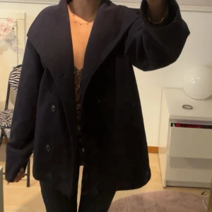 Marinblå kappa med bred krage - Säljer en svart kappa med bred krage och dubbelknäppning framtill. Jackan har en loose passform och långa ärmar, perfekt för dig som gillar en chill och stilren look. Materialet är tjockt och känns som ull, vilket gör den nice för kyliga dagar.