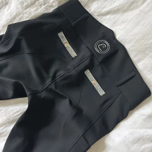 Svarta ridleggings Equest Dream - Snygga svarta ridleggings från Equest Dream med hög midja och silvriga detaljer vid fickorna. Byxorna har silikonprint på insidan av benen för extra grepp och logga tryckt på låret. Stretchigt material som sitter tajt och bekvämt.