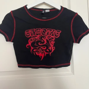 Svart croppad topp med röd ormmotiv - Svart croppad topp från hm med röda kontrastsömmar och tryck framtill med texten 'CHAOTIC' och ett detaljerat ormmotiv. T-shirten har korta ärmar och är tillverkad i mjuk bomull. Perfekt för dig som gillar edgy streetstyle.