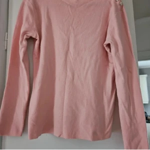 Ljusrosa långärmad tröja med knappar - Snygg ljusrosa långärmad tröja med rund hals och dekorativa knappar på ena axeln.