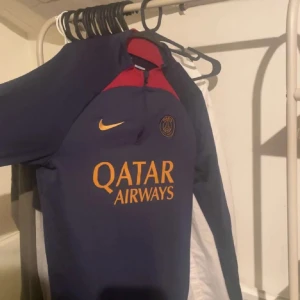 PSG långärmad fotbollströja Nike - Säljer en mörkblå långärmad fotbollströja från Nike med PSG-logga och Qatar Airways tryck i gult. Tröjan har röda detaljer vid halsen och är perfekt för dig som vill representera Paris Saint-Germain. Snygg och sportig design.