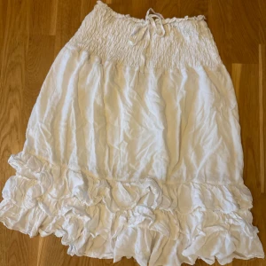 Vit volangkjol med smock och knyt - Supersöt vit kjol med smock i midjan och knytband. Kjolen har volangdetalj längst ner och är tillverkad i ett lätt tyg som känns perfekt för varmare dagar. Den har en luftig passform och slutar strax runt knäet.