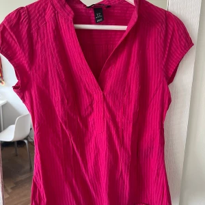 Rosa randig blus från H&M - Säljer en snygg rosa blus från H&M med diskret randigt mönster. Blusen har korta ärmar, v-ringad hals och lätt ståkrage. Materialet är mjukt och känns som bomull, perfekt för dig som gillar färg och enkel stil.