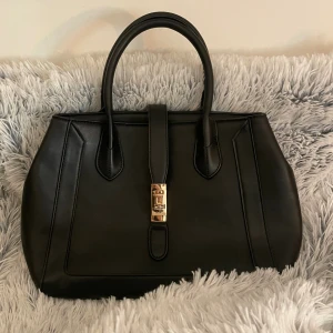 Svart handväska med gulddetalj - Stilren svart handväska i skinn med två handtag och snygg guldfärgad låsdetalj framtill. Väskan har en klassisk, strukturerad form och rymligt innerfack med mindre fickor. Perfekt för dig som gillar en clean och elegant look. Jättefint skick! 