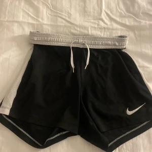Svarta Nike träningsshorts - Svarta träningsshorts från Nike med vita meshpaneler på sidorna och vit Nike logga på vänstra låret. Shortsen har elastisk midja med vit snörning. Perfekta för sport eller gympass.