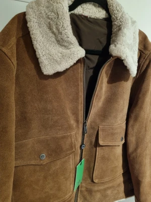 Mockajacka med teddykrage från La Martina - Snygg brun mockajacka från La Martina med fluffig teddykrage i beige. Jackan har två stora fickor med lock och dragkedja framtill. Insidan är fodrad och har en röd detaljficka. Broderat polomotiv på ryggen ger extra stilpoäng.