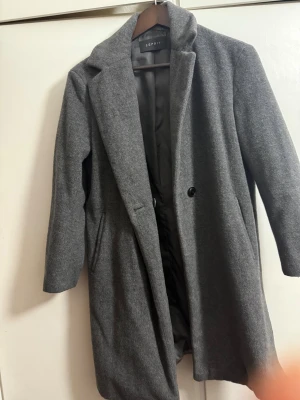 Grå kappa från Esprit i ullmix - Snygg grå kappa från Esprit i storlek S. Klassisk rak modell med krage, knäppning med en knapp och två sidofickor. Tillverkad i en mjuk ullmix som håller dig varm under kyliga dagar. Perfekt för dig som gillar stilrena och tidlösa plagg.
