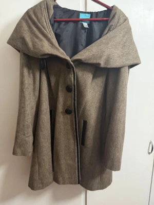 Beige kappa med stor krage från Andrée - Snygg beige kappa med stor, draperad krage och svarta knappar framtill. Jackan har en loose passform och är tillverkad i ullblandning med svart innerfoder. Perfekt för dig som gillar stilrena och klassiska plagg med en twist.