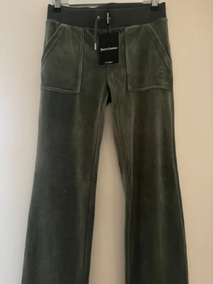 Gröna velourbyxor från Juicy Couture - Mörkgröna velourbyxor från Juicy Couture med bred resårmidja och snörning. Byxorna har stora fickor fram och bak, samt JC-logga broderad på ena fickan. Modellen är bootcut med mjuk och glansig finish, perfekt för en chill och trendig look.