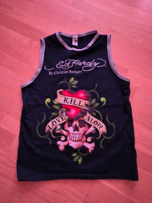 Ed Hardy marinblå linnetopp med tryck - Marinblå  linnetopp från Ed Hardy med grå kantband och stort färgstarkt tryck framtill med dödskalle, hjärta och texten 'Love Kills Slow'. Toppen har ärmlös design och logotyp både fram och bak. Materialet känns som mjuk bomull.