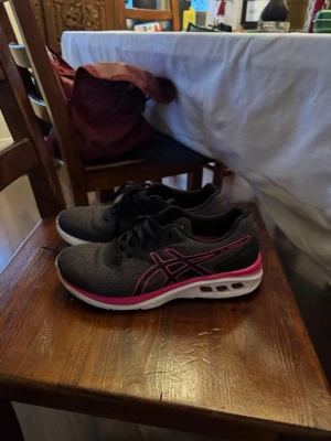 Svarta och rosa Asics sneakers - Säljer ett par snygga Asics sneakers i svart med rosa detaljer och vit sula. Skorna har snörning och är tillverkade i syntetmaterial med gummisula. Perfekta för träning eller en aktiv vardag. Klassisk Asics-logga på sidan och bekväm passform.