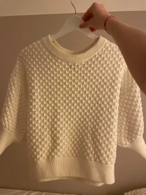 Vit stickad tröja med strukturmönster - Säljer en vit stickad tröja med snyggt strukturmönster och ribbade muddar vid hals, ärmslut och nederkant. Tröjan har en rund halsringning och korta ärmar, perfekt för lager-på-lager och chill stil.