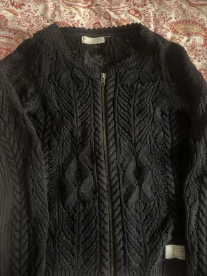 Odd Molly kofta  - Svart stickad cardigan jacka från Odd Molly med grova flätstickade detaljer och dragkedja framtill. Jackan har långärmade ribbade muddar och ett märke med text på ärmen. Perfekt för dig som gillar snygga och mysiga lager. Passar s 