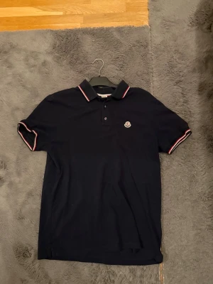 Marinblå Moncler pikétröja  - Säljer en marinblå pikétröja från Moncler i storlek L. Tröjan har klassisk krage med två knappar och vita samt röda detaljer på krage och ärmslut. Moncler-logga på bröstet. Tillverkad i bomull för en skön känsla. OBS storleken är L men kan funka på dig som bär M. 